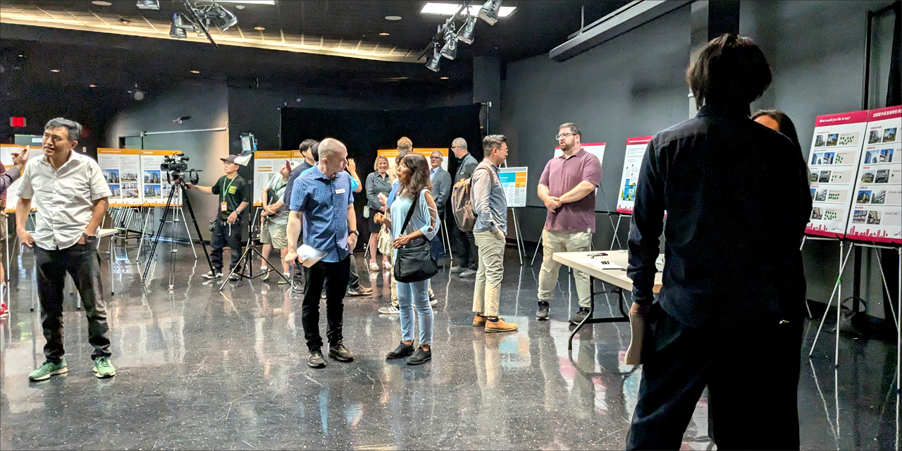 Wide angle shot of people standing indoors for an information open house. They are talking to other or looking at display boards.
