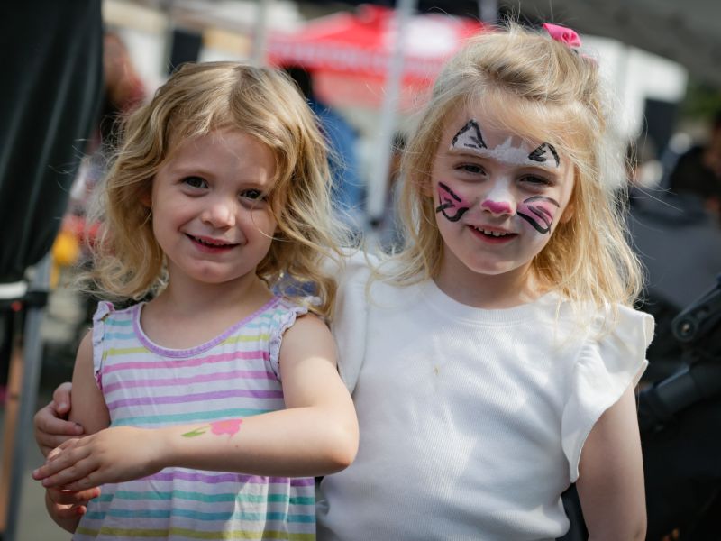 two female children at a community event, one with her face painted like a cat, the other with a flower painted on her arm